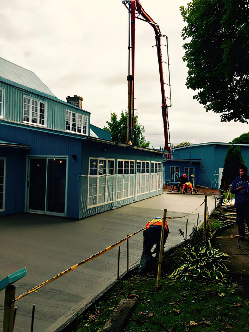 Une pompe à béton déployée au-dessus d'un bâtiment communautaire bleu, deux ouvriers finissent la dalle fraîche