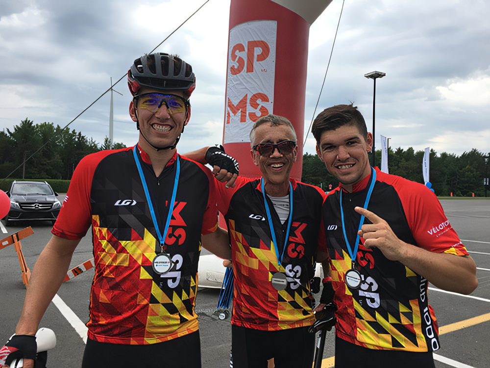 Trois cyclistes souriants en maillot rouge, or et noir, médaille au cou, devant une arche gonflable