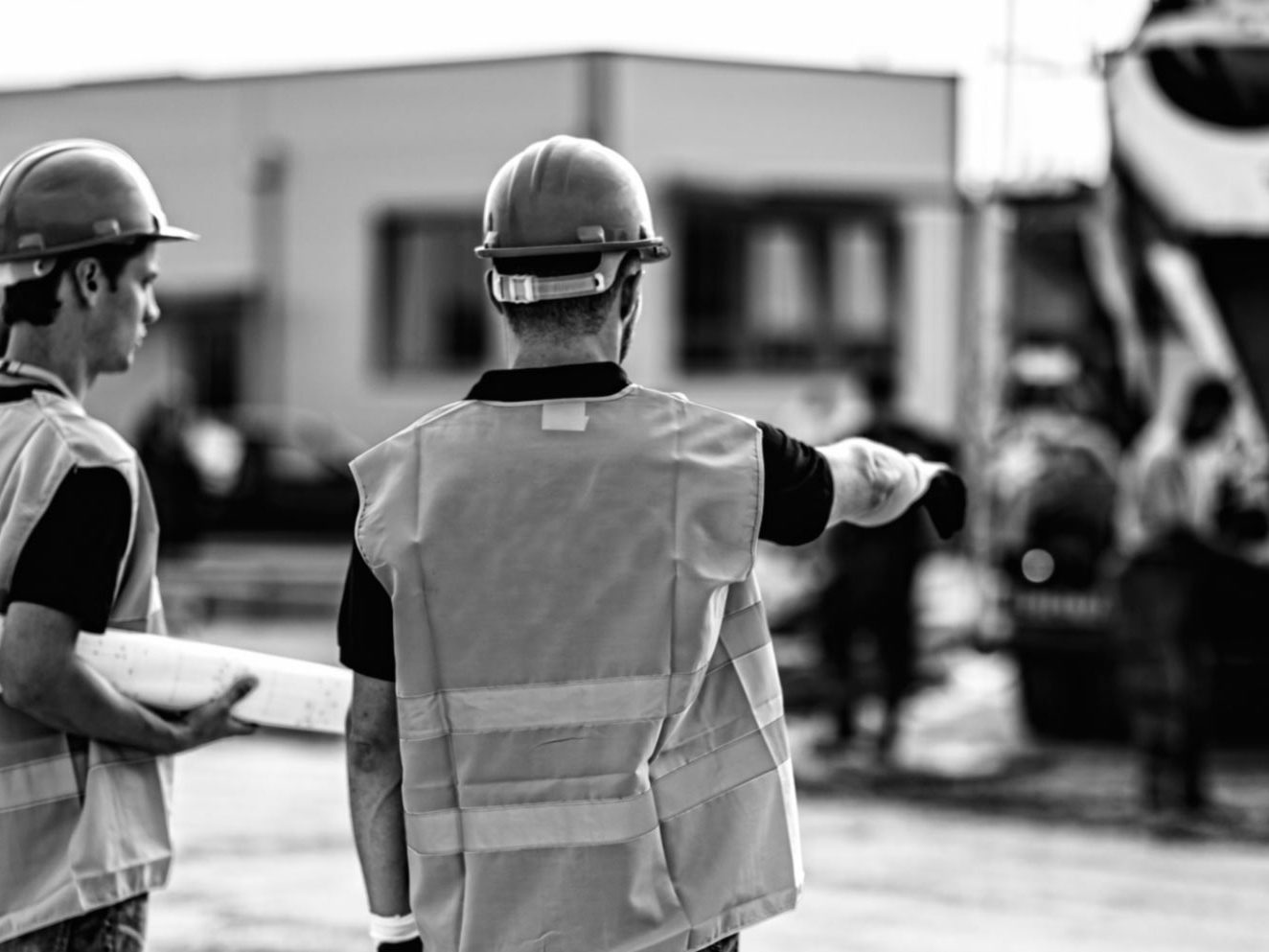 Deux contremaîtres en casque et dossard discutent sur un chantier, une bétonnière derrière eux, photo en noir et blanc